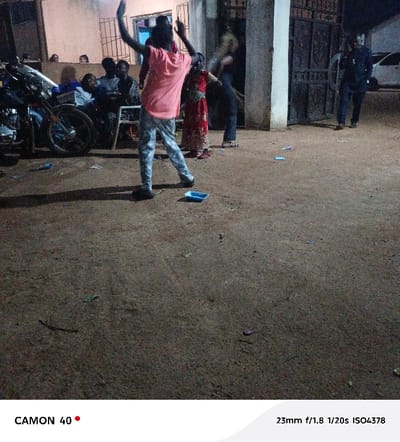 The image captures an outdoor evening scene in Egede, Nigeria, illuminated by artificial light. In the foreground, a person stands with their back to the camera, wearing a pink short-sleeved shirt and light-colored patterned trousers. Their arms are raised overhead, and their legs are spread, indicating a dynamic posture. A small, light blue plastic container rests on the unpaved, reddish-brown dirt ground directly in front of this individual. Scattered debris, including small blue and reddish fragments, is visible across the ground.

To the immediate right of the central figure, a smaller individual wearing a red patterned dress is partially visible, also in motion. Further right, another person in dark clothing is partially obscured. On the far left, a black motorcycle with chrome components is parked. Behind the motorcycle and against a light-colored building wall, a group of approximately 5-7 individuals is seated on chairs or benches. Additional figures are partially visible further left, behind a wall. On the right side of the frame, a dark metal gate with decorative patterns is open, through which two figures are seen walking away from the camera. A white vehicle is partially visible beyond the gate in the far background. The scene is characterized by warm artificial lighting that casts distinct shadows, notably a long shadow from the central figure extending towards the bottom left.