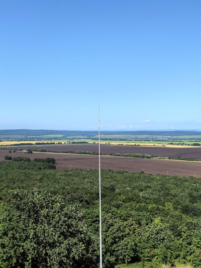 This is a wide-angle, landscape photograph taken from an elevated position overlooking a rural area. The foreground is dominated by a dense, dark green forest. A tall, slender, metallic pole, likely an antenna or lightning rod, is positioned vertically in the center of the frame, extending from the bottom edge to near the top.
Beyond the forest, the landscape opens up to a series of agricultural fields. Closest to the forest is a field of dark, tilled soil. Further in the distance, there are fields of golden crops, possibly wheat or barley, and patches of green vegetation. A treeline separates these fields.
In the far distance, rolling hills covered in dark green vegetation form the horizon. The sky is a clear, bright blue with no clouds visible. The image appears to be taken on a sunny day, with no people or animals visible in the frame. The location is identified as Akhmerovo, Russia.