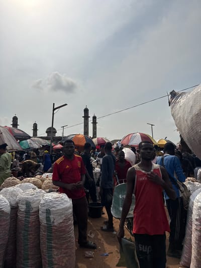 The image depicts a bustling outdoor market in Ogere, Nigeria, under a partly cloudy sky. The foreground is dominated by numerous large, stacked, translucent white sacks filled with a reddish-brown produce, consistent with onions. Two adult males are prominent in the immediate foreground, both looking towards the camera. The man on the left wears a red short-sleeved shirt with a yellow crest and dark pants; the man on the right wears a red tank top and dark shorts, holding a piece of dark fabric, with a green metal wheelbarrow beside him.
The midground and background feature a dense crowd of people, many engaged in market activities such as moving goods, standing, or attending to stalls. Various market stalls are visible, shaded by umbrellas and awnings in colors including red and white stripes, orange, and green. In the distant background, several tall, light-colored mosque minarets with conical tops are discernible against the sky, alongside what appears to be a dome structure. A black street light pole with a horizontal arm and a single lamp is positioned near the center-left. Overhead lines traverse the upper portion of the frame. The ground throughout the market area is unpaved dirt.