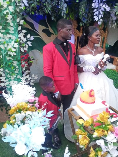 An event scene in Amokvo, Nigeria, displays three individuals standing amidst artificial foliage and floral decorations. An adult male on the left, with dark skin and short dark hair, wears a red blazer with black lapels, a black shirt with a decorative bolo tie, and black trousers. He stands with his body angled slightly right, looking towards the right. To his right, an adult female with dark skin, dark hair pulled back with a black headband, wears a white off-the-shoulder wedding gown, white lace fingerless gloves, a prominent silver-colored necklace, and earrings. She holds a white, cylindrical object, possibly a microphone or small fan, in her gloved right hand and has a small black bag on her left shoulder. She also looks towards the right. Between the adult male and a tiered cake, a young boy with dark skin and short dark hair wears a matching red blazer with black lapels, a black shirt, and black trousers. He holds a clear plastic bottle in his left hand and also looks towards the right.
In the foreground, a two-tiered cake rests on a wooden-framed stand. The bottom tier is white and rectangular, while the top tier is an irregular, yellowish-orange, stylized shape with red decorative elements. A blue marker or pen is inserted into the top tier. Numerous artificial flower arrangements in white, light blue, yellow, pink, and red are arranged around the base of the cake stand and in the immediate foreground. The ground is covered in artificial green turf. The background features artificial green and white foliage, a partial view of an ornate gold-colored chair, and a green backdrop with abstract designs, suggesting an indoor or decorated outdoor setting for a formal event.