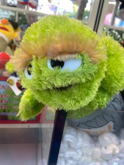 The image shows a close-up of a fuzzy green stuffed toy, likely a puppet, with blonde hair. It has large white eyes with black pupils and a small mouth. The puppet is inside a clear plastic enclosure, possibly a prize machine. In the background, blurred figures and colorful objects are visible, suggesting an indoor environment like an arcade or amusement center. A portion of a box with text is visible on the left side of the image, but the text is mostly unreadable. The toy is mounted on a black rod. The scene appears to be located within the city of Baunatal, Germany.