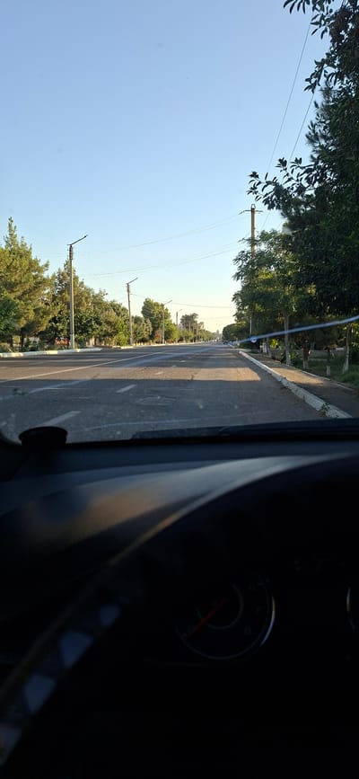 The image captures a roadside view in Kogon City, Uzbekistan, likely taken from the interior of a vehicle. The perspective is from the right side of the car, looking forward and slightly to the left. The scene is dominated by a paved road stretching into the distance, bordered by a sidewalk and a line of trees. Electric poles with wires are visible along the roadside, suggesting an urban or semi-urban setting.
The time of day appears to be late afternoon or early evening, with long shadows cast across the road by the sun, which is positioned low in the sky. The sky is clear and bright blue. The vegetation consists of leafy trees, some appearing to be deciduous, interspersed with possibly some evergreen varieties.
In the foreground on the right, a dark, indistinct object, likely part of the car's interior, partially obscures the view. A circular gauge with a red needle, possibly a speedometer or fuel gauge, is dimly visible within this dark area.
The road is marked with white lines, and some parked vehicles can be seen further down the road, indicating normal traffic activity. The overall atmosphere is calm and typical of a residential or commercial street. There is no visible text in the image.