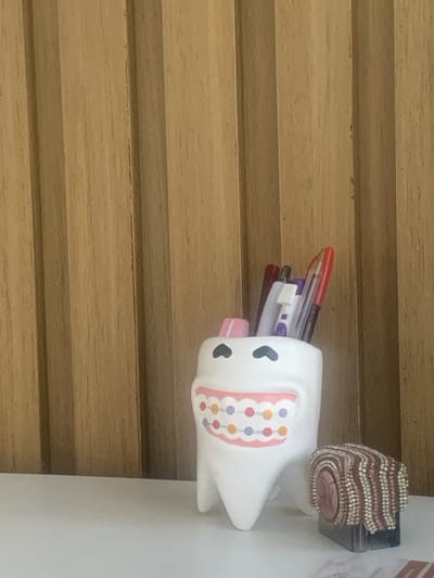 The image displays an indoor scene situated in Lauro de Freitas, Brazil. A white, ceramic, tooth-shaped container, approximately 15 cm in height, stands on a white, flat surface. The container is designed with a cartoonish face, featuring a wide pink mouth with colorful orthodontic braces depicted as multiple circular dots (orange, purple, and pink). Two small, black heart-shaped outlines are present above the mouth, serving as eyes. The container holds multiple writing instruments, including pens and pencils of various colors (e.g., red, white, purple). To the immediate right of the tooth-shaped container, a dark, rectangular object, possibly a stamp or ink pad, is partially visible. It is topped with a textured cover composed of interwoven or beaded strands in shades of brown, pink, and white. This cover features a central circular design with the letters "DR" inscribed. The background consists of vertical light brown wooden paneling with visible grain. No individuals are present in the scene. The environment appears well-lit by ambient indoor light.