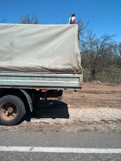 A side view of a weathered truck with a light-colored tarp-covered cargo bed is captured against a bright blue sky. The truck's rear wheel, with a rusty rim, is visible in the foreground. The background shows a dry, brush-filled landscape with bare trees, suggesting a rural or undeveloped area. A white stripe painted on an asphalt road runs along the bottom of the frame. The image appears to have been taken on a sunny day.