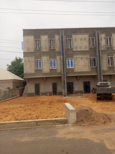 This media file depicts an outdoor scene in Baccure Kofare, Nigeria, likely during the daytime under overcast skies. The primary subject is a multi-story building under construction. The building's facade is rough, unfinished concrete with recessed square sections above and below the windows. It features several windows on multiple levels, some with white bars and others with blue-tinted glass. Some windows are open, while others are closed.

In front of the building, there is a large area covered in reddish-brown soil, indicative of a construction site. A concrete curb separates this area from a paved road in the foreground. To the left of the building, a smaller structure with a light-colored, pitched roof is partially visible. Piles of building materials, possibly stones or bricks, are stacked near this smaller structure.

To the right of the building, a light-colored truck with a wooden flatbed is parked on the soil. A person wearing a red garment is seated on the ground near the truck. A set of concrete stairs leads up to one of the building's entrances. There are also several vertical pipes running along the side of the building. Some faint graffiti or markings, appearing to be numbers, are visible on the wall near one of the ground-floor entrances. The overall impression is one of ongoing construction and development in a somewhat rural or semi-urban setting.
