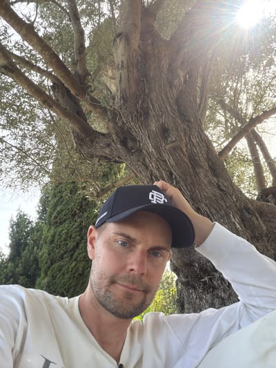 A man with light skin, blue eyes, and short brown hair, wearing a white long-sleeved shirt and a black baseball cap, is photographed outdoors in what appears to be Montuïri, Spain. The man is looking towards the camera, with his left hand resting on the brim of his cap. He has a light beard and is positioned in the lower right quadrant of the image. Behind him, a large tree with thick branches and green leaves dominates the background. The sun is visible in the upper right corner, casting bright rays through the tree. To the left of the tree, there is a dense line of green evergreen trees. The overall setting suggests a sunny day in a natural environment.