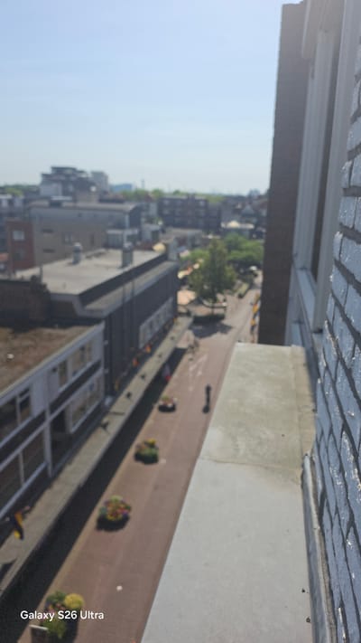 The image provides an aerial view of a street in Vlaardingen, Netherlands, looking downwards and to the left. The scene depicts a city street lined with buildings of varying architectural styles, some appearing to be commercial with awnings and windows. Several planters with colorful flowers are arranged along the side of the street. A few individuals are visible on the street, one walking and another appearing to be stationary. The sky is clear and blue. The foreground shows a concrete ledge and the brick facade of a building, suggesting the photo was taken from an elevated position. The text "Galaxy S26 Ultra" is visible in the bottom left corner, indicating the device used to capture the image.