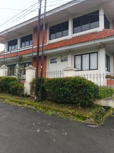 An exterior daytime photograph captures a two-story building in Masaiti, Zambia, under an overcast sky. The structure displays white painted walls, a section of reddish-brown brick on the left facade, and a terracotta-tiled roof with prominent white eaves. Both floors feature numerous dark-framed windows; ground-floor windows on the right are equipped with white security grilles. A black wrought-iron fence with 'X' patterns and concrete pillars borders the property, fronted by a dense green hedge. A dark asphalt road, marked with light stains, occupies the foreground, separated from the property by a moss-covered concrete curb. Two dark utility poles with overhead wires are positioned centrally in front of the building. A planter containing reddish-orange flowers is visible behind the fence on the far right. No individuals are present in the scene.