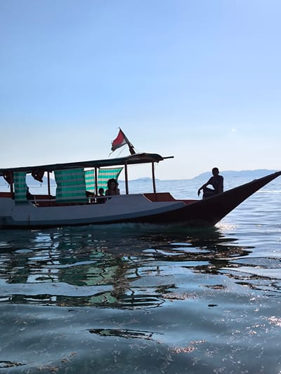 A long, narrow boat with a canopy is shown sailing on the water, possibly near Tanjung Boleng, Indonesia. The boat has a white hull with an orange trim, and green and white striped curtains hang from the canopy. A red and white flag flies from a pole at the back of the boat. In the background, a person is seated on the bow of the boat, silhouetted against the sky. Several other people are visible inside the boat, one of whom appears to be a woman with curly hair looking to the right. The water is choppy, reflecting the sky and the boat. The scene appears to be set on a clear, sunny day with distant hills or islands visible on the horizon.