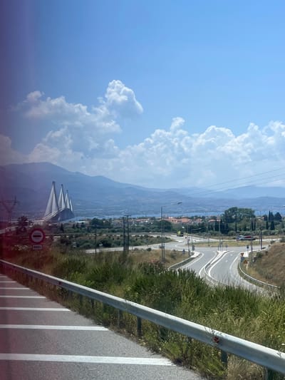 The image depicts a scenic view of the Rio–Antirrio Bridge in the Municipal Unit of Antirrio, Greece. The foreground shows a paved road with white lane markings and a metal guardrail, bordered by dry grass and vegetation. In the mid-ground, a complex highway interchange with multiple lanes and road signs is visible. Beyond the interchange, the distinctive cable-stayed Rio–Antirrio Bridge spans across a body of water, with its tall pylons prominently displayed. The background features rolling hills and mountains, a coastal town with red-tiled roofs, and a vast expanse of blue sky filled with fluffy white clouds. The overall impression is a sunny, clear day in a Mediterranean landscape.