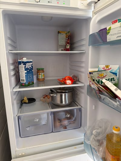 This is a medium shot, eye-level, indoor, close-up photograph of the inside of a white refrigerator. The refrigerator is mostly empty, with a few items on the shelves and in the door compartments. On the top shelf, there is a package of red tomatoes with green stems and a small jar of what appears to be baby food. On the middle shelf, there is a carton of milk with a blue and white label, a jar of baby food with a green lid, and a small black dish with a piece of yellow food on it. There is also a pot with a lid next to a plastic bag containing some food items. The door of the refrigerator has several compartments containing various packages of food items. In the bottom compartment of the refrigerator, there are two drawers. To the right of the refrigerator, there is a bottle of cooking oil. The lighting is bright and even. The refrigerator appears to be clean and well-maintained. The city of Verkhnevilyuysk, Russia, is known for its extremely cold climate, with average January temperatures around -40 degrees Celsius. This may explain why there are few perishable items in the refrigerator.