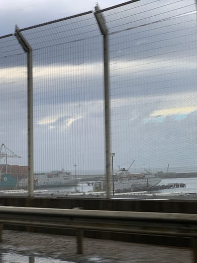 This image depicts a port in Ksar El Majaz, Morocco, viewed through a prominent wire mesh security fence and a dark metal guardrail. The foreground shows a wet, dark ground surface. An overcast sky with patches of brighter, diffused light dominates the upper portion. In the midground, within the harbor, two large white multi-deck vessels are docked. The vessel on the left bears the partially visible name "BALEARIA" on its hull. The vessel on the right, equipped with multiple red cranes on its upper decks, is moored at a pier. Additional port infrastructure, including a large green gantry crane, shipping containers, industrial buildings, and light poles, is visible within the harbor area. No individuals are discernible. The scene portrays a stationary port environment under an overcast sky.