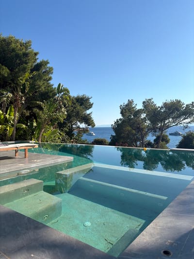 The image depicts an infinity pool overlooking the sea. The pool has a turquoise-colored tiled bottom and concrete steps leading into it. There are several small fish swimming in the shallow steps of the pool. A wooden lounge chair is visible on the tiled edge of the pool. Lush green trees and foliage surround the pool area, with palm trees visible on the left. In the background, the blue sea stretches out to the horizon, dotted with a few white yachts. The sky is clear and bright blue. The scene appears to be a luxurious vacation setting, likely in the city of Vouliagmeni Municipal Unit, Greece, given the coastal environment and Mediterranean vegetation.