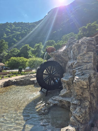 The image depicts a water wheel situated within a rocky, man-made cascade. Water flows from an unseen source above, spilling over the rocks and onto the black spokes of the wheel, creating a dynamic spray of water. The wheel is positioned in a pool of shallow, clear water that reflects the sunlight, causing shimmering patterns on the surface.

Behind the water feature, a lush green mountainside covered in trees slopes upwards towards a bright blue sky. Sunlight streams from the left side of the frame, casting distinct rays and creating lens flare. The overall scene suggests a pleasant, sunny day, likely in the late morning or early afternoon given the angle of the sun.

In the foreground, to the right of the water wheel, a paved walkway is visible, lined with rough-hewn rocks that form the edge of the water channel. Beyond the walkway, there are glimpses of park-like landscaping with benches and trees, and a road with a parked car. A brown, ceramic-like jug is partially visible at the top left, from which water appears to be pouring. The scene evokes a sense of natural beauty enhanced by man-made elements, likely a tourist attraction or a decorative feature in a park or garden. No visible text is present in the image.
