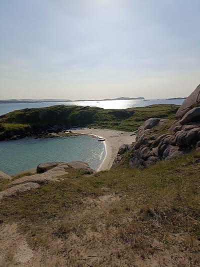 This is a photograph of a small, secluded cove on the coast of Carnboy, Ireland, under a clear sky. The foreground features dry, sandy ground with sparse, brown vegetation and large, textured grey boulders. The middle ground reveals a crescent-shaped sandy beach meeting calm, turquoise water. Several small boats, including what appear to be kayaks, are beached on the sand. A few individuals are visible on the beach. The background consists of a low, green, grassy island with rocky outcrops, and beyond that, the expansive, shimmering sea under a bright sun, with distant islands silhouetted against the horizon. The overall impression is of a peaceful, natural coastal landscape.