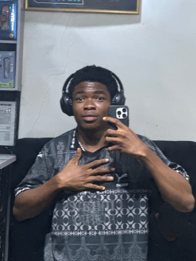 A young man is taking a selfie with his phone. He has headphones on and is wearing a patterned shirt. He appears to be sitting. The location is likely indoors in Ogwashi-Uku, Nigeria, based on the visual style and context of the picture.