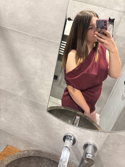 A young woman is taking a mirror selfie in a modern bathroom. She is wearing a dark red, off-the-shoulder dress and holding a phone with a colorful case in her right hand. The bathroom features large, light gray tiles on the walls and a stone sink with chrome faucets. A towel rack is visible in the background. The image is taken from a slightly high angle, looking down at the woman and the sink.