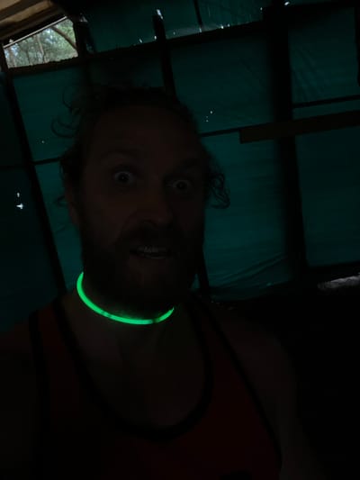 The image is a close-up, self-taken photograph of a man inside a dark structure. The man has a beard and wild, curly hair. His eyes are wide open, and his mouth is slightly ajar, revealing his teeth. He is wearing a neon green glow necklace that is brightly illuminated. The background appears to be some sort of makeshift shelter, with a greenish tarp-like material covering the walls and ceiling, and wooden beams supporting the structure. Glimpses of trees and the sky are visible through an opening at the top left of the frame. The overall lighting is dim, with the glow necklace providing the primary light source for the foreground. The location is described as being in the city of Luzhki, Russia.