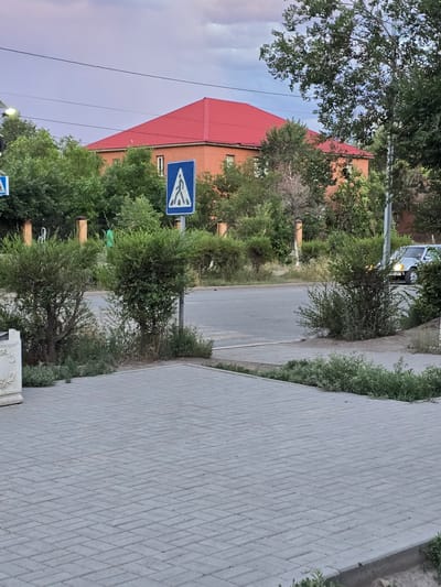 The image depicts a street scene in Satbayev, Kazakhstan. A sidewalk paved with gray bricks runs diagonally from the lower right to the upper left of the frame. Green bushes and trees line the edge of the sidewalk and the street. A red-roofed building, likely an apartment block, is visible on the left side of the street. A blue traffic sign with a white pedestrian symbol is mounted on a pole. In the background, a silver car is parked by the side of the road, and power lines crisscross the sky. The sky is a pale purple and blue, suggesting it might be early morning or late evening. The overall mood of the image is calm and ordinary, capturing a slice of everyday life in a residential area.