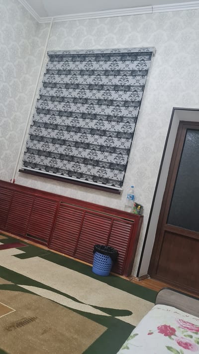 A domestic interior scene, likely a bedroom or living area, is depicted from a slightly elevated perspective. The room features a patterned wallpaper with a damask motif in light grey and white. A horizontal blind with alternating opaque and sheer stripes, also featuring a dark floral pattern, is drawn across a window. Below the blind, a long, dark red wooden cabinet with louvered doors spans the lower portion of the wall. To the right of the cabinet, a blue plastic trash can with a black liner is visible. Further to the right, a portion of a wooden door with frosted glass is present. In the foreground, a carpet with a geometric design in cream, green, and brown tones covers the floor. A bottle of water and a small packaged item are on top of the cabinet. The scene is static and appears to be a still photograph. The location is identified as Karshi City, Uzbekistan.