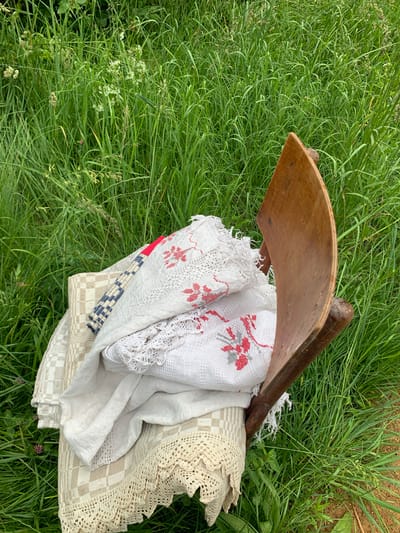 An old wooden chair with a curved back is placed on a patch of tall green grass in an outdoor setting. The chair is draped with several folded pieces of fabric, including what appears to be a white embroidered tablecloth with red floral patterns and another fabric with a checkered design and lace trim. The surrounding environment is dominated by lush, verdant grass, with some wildflowers interspersed. The lighting suggests it is daytime, possibly late afternoon, with soft light illuminating the scene. The image appears to have been taken at an eye-level perspective, looking down slightly at the chair and its contents.