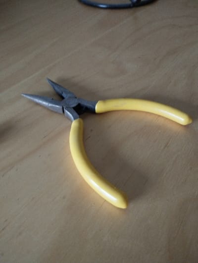 A pair of long-nose pliers with yellow plastic-coated handles and grey metallic jaws lies angled on a light-toned wooden surface with visible grain patterns. The pliers are in an open position, with the handles spread apart and the tapered jaws separated, pointing towards the upper left. Illumination from the upper left casts a shadow of the tool towards the lower right. A dark, curved object, out of focus, is partially visible in the upper background. The scene is located in Fontenay-sous-Bois, France.