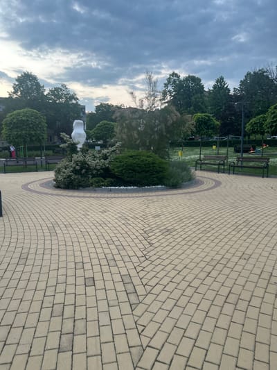 The image captures a peaceful public park or town square in Dzhebel, Bulgaria, under an overcast sky. A significant portion of the foreground and midground is dominated by a meticulously paved pathway made of light beige rectangular pavers. This pathway features an intricate pattern, including a large semi-circular section bordered by darker, reddish-brown pavers, which outlines a landscaped green island.
Within this landscaped island, various shrubs and bushes are visible, some neatly manicured, others with white blossoms. A prominent, abstract white sculpture, with smooth, organic curves, stands among the greenery, serving as a focal point.
Beyond the paved area and the central island, a well-maintained green lawn stretches out, featuring several dark wooden park benches with metal supports, inviting visitors to rest. Further back, numerous trees with dense green foliage, including both deciduous and coniferous types, form a lush backdrop, partially obscuring the distant horizon and any potential buildings. The cloudy sky, with its soft, diffused light, suggests either early morning or late afternoon. The scene is devoid of people, contributing to an overall tranquil and serene atmosphere. No visible text is present in the image.
