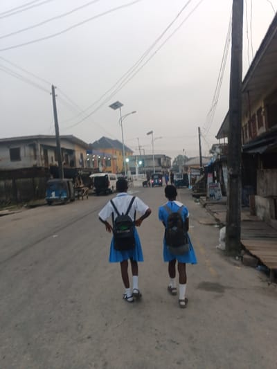 Two schoolgirls in blue uniforms and backpacks walk down a paved road away from the camera. Various buildings line the street, including some with windows and a blue and orange building in the distance. Streetlights illuminate the road. Several auto rickshaws are parked and moving ahead of the girls. Overhead power lines run across the sky. The sky appears overcast.