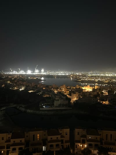 This is a night time aerial view of Al Jazeerah Heritage Village, United Arab Emirates. The scene is illuminated by artificial lights from buildings, streetlights, and construction sites in the distance. In the foreground, there are several multi-story residential buildings with terracotta roofs and illuminated windows. A dark body of water, likely a canal or inlet, runs through the middle ground, reflecting the city lights. Beyond the water, a densely populated area of similar residential buildings stretches towards the horizon. In the far distance, a large industrial or construction area is visible, characterized by numerous bright lights and tall cranes. The sky is dark and overcast, with a subtle gradient of light from the city below. There are no people or animals visible in the frame. The overall impression is one of a vibrant, active urban environment at night.