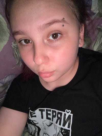 The image displays a close-up portrait of a person lying down, viewed from an overhead perspective. The subject appears to be young, with light-to-medium skin, dark brown eyes, and dark hair. Their head rests on a patterned surface, possibly bedding or a pillow, with hues of muted green and purple.

Notable features include two piercings on the person's face: a horizontal eyebrow piercing with two silver studs on their left eyebrow, and a silver septum ring in their nose. The person is looking directly towards the camera with a calm, neutral, or slightly contemplative expression. Their lips are naturally full and slightly parted.

They are wearing a black short-sleeved t-shirt adorned with a white graphic design. A portion of the text on the shirt is visible and appears inverted, reading "ГЕРЯЙ" (Geryay in Cyrillic script). The graphic itself seems to be an illustrative style, though largely cropped, showing parts of what could be a mythical creature or figure.

The lighting suggests an indoor setting, likely somewhat dim, with the person's face well-lit. The image is a focused shot on the person's upper body and face, capturing a casual and intimate moment. There are no explicit indicators of time of day or weather.