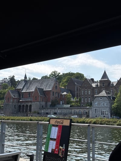 The image depicts a waterfront view in Dinant, Belgium, likely from a cafe or restaurant terrace, as indicated by the presence of a menu stand in the foreground. The menu features the Coca-Cola logo prominently, along with Italian-themed dishes, suggesting a dining establishment. Beyond the water, a collection of historic buildings, characteristic of Dinant's architecture, line the bank. The most prominent structure is a large, red-brick building with arched ground-level openings and a tall, spire-topped tower, which appears to be a former abbey or church. Other buildings in the background showcase a mix of architectural styles, with grey stone facades and numerous windows. The water's surface is rippled, reflecting the sky, which is partly cloudy. Greenery, including trees and bushes, surrounds the buildings and extends up the hillside. A metal railing separates the foreground terrace from the water. The scene is bathed in daylight, suggesting it was taken during the daytime. There are no people clearly visible in the main body of the image, though a few figures might be seen in the distance near the buildings along the riverbank. The overall impression is one of a tranquil, scenic European town.