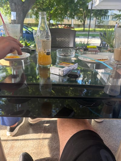 The image depicts an outdoor table at a cafe in Dalgopol, Bulgaria. On the glass tabletop, there is a bottle of orange juice, a glass with a red straw, a cigarette pack, a lighter, an ashtray with a cigarette, and two glasses of iced coffee. The legs and feet of two people are visible, one wearing black sneakers with white soles and blue shorts, and the other wearing black athletic shoes and black shorts. In the background, trees and a building can be seen, suggesting a sunny day. The atmosphere appears casual and relaxed, as if the individuals are enjoying a break.