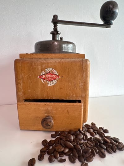 This media file features a close-up, overhead view of an antique wooden coffee grinder situated on a white surface, likely a wall or tabletop, in Lapės, Lithuania. A pile of roasted coffee beans spills out from the right side of the grinder, suggesting it has recently been used or is about to be. The grinder itself is primarily made of light-colored wood, with a metal grinding mechanism and a dark wooden handle. A small, round knob made of wood is visible on the front of the grinder, likely for adjusting the grind coarseness.
A prominent detail on the side of the wooden grinder is a torn, circular red and white sticker with text. The visible text reads "LEN BROCK'S" and "DEAL," possibly indicating the brand or model of the coffee grinder. The lighting appears to be even, with no distinct shadows to indicate a specific time of day, and the white background suggests an indoor setting, free from weather considerations. The overall impression is one of vintage domesticity and the simple act of preparing coffee.