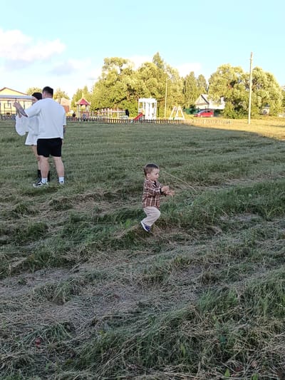 In Smolino, Russia, on a grassy field under a clear sky with scattered clouds, two adults and a young child are present. An adult male, wearing a white t-shirt and black shorts, stands with his back to the camera, his left arm raised. Beside him, an adult female, also in a white outfit, faces him, holding a phone in her right hand and gesturing with her left. In the mid-ground, a young boy in a brown plaid shirt and tan pants runs towards the right side of the frame, his arms outstretched. Behind them, a wooden fence encloses a playground area with swings and colorful structures. Trees and houses are visible in the background.