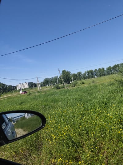 The image is a photograph taken from inside a vehicle, looking out towards a rural or suburban landscape under a clear blue sky. In the foreground, a side-view mirror reflects a dirt road and some low-lying structures, possibly garages. Beyond the mirror, a vast expanse of green grass, dotted with small yellow flowers, stretches towards a treeline in the middle ground. Several utility poles with electrical wires are visible, suggesting the presence of infrastructure. In the distance, a large, multi-story residential building stands, with a red car parked in front of it. The overall scene suggests a sunny day in a relatively undeveloped area on the outskirts of a town or city.