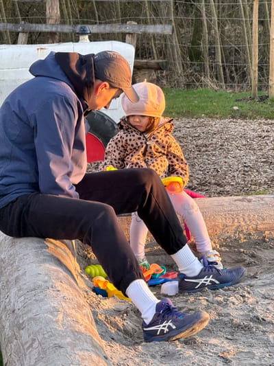 In Olingen, Luxembourg, an outdoor scene depicts an adult and a child in warm, low-angle sunlight. An adult male, wearing a dark blue hooded jacket, black sweatpants, a dark baseball cap, and dark blue ASICS sneakers with white socks, sits on a large, weathered wooden log. He is positioned facing right, looking downwards towards a young child. The child, dressed in a light pink bonnet, a beige and black leopard-print jacket, and light pink leggings, sits on the sandy ground to the adult's right, looking downwards. Several brightly colored plastic sand toys, including shovels and buckets, are scattered on the sand around both individuals. In the background, to the left, a portion of a white object with a red wheel, possibly a tractor, is visible. Behind the subjects, a rustic fence composed of horizontal wooden planks and wire runs across the frame, with a dense area of slender, bare trees or saplings beyond it. The ground consists of light-colored sand or dirt.