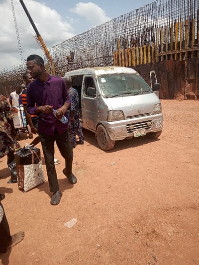 The scene takes place in a dusty, open-air area, likely a construction site in Otta, Nigeria, as indicated by the coordinates. A silver minivan is parked in the center of the scene, facing the viewer. Several people are present, with one man in a purple shirt standing prominently to the left of the van. He is holding a water bottle and appears to be looking at something in the distance. Other individuals, both men and women, are situated in the background, some holding bags and boxes. A large, rust-colored metal fence encloses the site, indicating ongoing construction.  The presence of a yellow crane in the background further suggests that the location is an active construction zone. The ground is dry and covered with reddish-brown dirt, typical of the environment in this area. The overall atmosphere seems casual and daily, as if the people are involved in a routine activity.