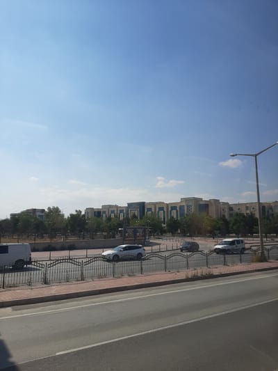 The image captures a street scene in Karatay, Turkey, under a clear blue sky with scattered white clouds. In the background, a large, modern building complex is visible, featuring a repeating architectural pattern of blue-tiled sections and beige facade elements. The building appears to be an official or institutional structure, as indicated by the partial text "KONYA ADALET SARAYI" (Konya Courthouse) visible on a sign.
In the middle ground, a road is lined with a black wrought-iron fence and a red brick sidewalk. Several vehicles are present on the road. A white van is parked on the left side of the road, and a white sedan is driving on the right side of the road. Further down the road, a dark-colored car and a white SUV are also visible. Trees and greenery are interspersed behind the fence, suggesting a landscaped area or park. A tall streetlight stands on the right side of the image.
The foreground shows a section of asphalt road, indicating the perspective is from another vehicle or roadside. The overall impression is a well-maintained urban environment on a bright, sunny day.
