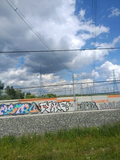 The image captures a scene of electrical transmission towers and power lines against a sky with a mix of blue sky and dramatic clouds. In the foreground, a section of a wall is covered in colorful graffiti. The ground in front of the wall is covered with gray gravel, and beyond that, lush green grass and vegetation are visible. The overall impression is one of an industrial or semi-industrial area with signs of urban art. The presence of the electrical infrastructure suggests proximity to power distribution networks. The location is identified as Vyshnyakovo, Russia.
