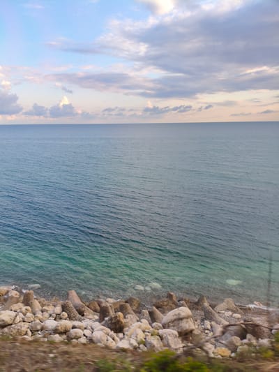 The image depicts a coastal scene in Chigripshi, Abkhazia. The horizon separates a vast expanse of the Black Sea from a cloudy sky. The sea is a deep blue, transitioning to a lighter, turquoise hue near the shore. Along the coastline, a collection of large, irregularly shaped rocks and concrete tetrapods are visible, serving as a breakwater. The foreground shows a narrow strip of earth with sparse vegetation. The overall impression is one of a natural, somewhat rugged coastline.