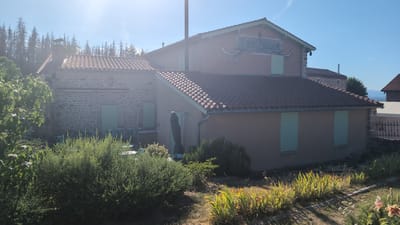 The image shows a building with a sign that reads "Ess'Folie Cafe" located in Chambles, France. The building has a terracotta tiled roof and is partially covered in stucco and stone. It has light green shutters on the windows. There is vegetation, including trees and bushes, in front of the building. The sky is clear and blue. The sun is shining brightly, creating a lens flare effect in the upper left corner of the image. The scene appears to be an outdoor seating area or a cafe setting.