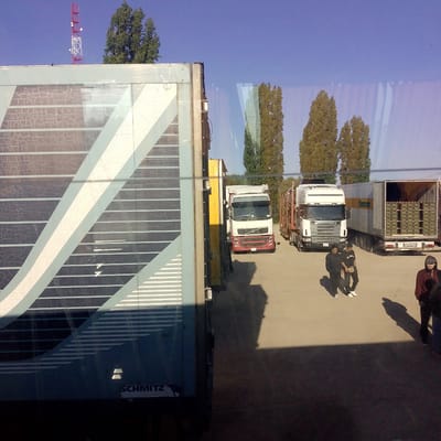 This is a daytime, eye-level, wide-angle photograph taken from inside a vehicle, looking out at a parking lot with several trucks. The foreground is dominated by the side of a large, blue and white striped trailer, which appears to be a Schmitz trailer based on the visible logo. The trailer has a weathered, cracked surface.

In the middle ground, there are several semi-trucks. One white Volvo truck with a red grille is parked facing left. To its right, another white semi-truck with a trailer filled with cases of bottled beverages is parked. The trailer is open at the back, revealing the stacked cargo. Two men are walking in the parking lot between these trucks, one wearing a black hooded sweatshirt and dark pants, the other in a black t-shirt, dark pants, and a fanny pack. Further to the right, a person wearing a dark hooded jacket and dark pants stands partially obscured by shadow.

In the background, there are several tall trees with green foliage. A red and white telecommunications tower is visible against a clear blue sky. The overall scene suggests a logistics or transportation hub. The image does not provide specific visual cues to confirm the location within Vetyutnev, Russia, but it depicts a typical scene at a truck stop or industrial area.