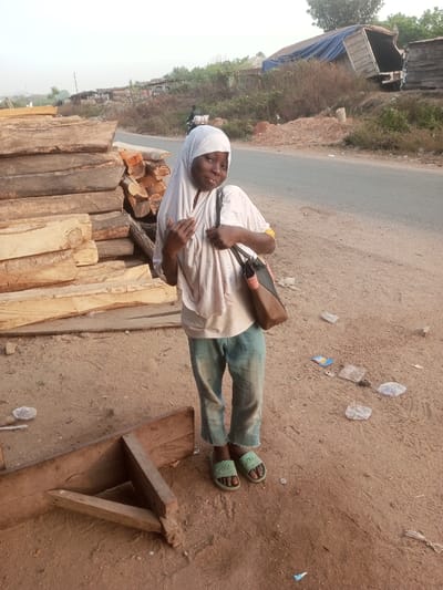 A young Black person stands posing for the camera on a dusty roadside in Ebiya, Nigeria. They are wearing a white hijab, a light-colored patterned long-sleeved top, light blue cropped jeans, and light green slides with a visible text or logo on them. A brown and black shoulder bag hangs over their left shoulder. The person has a subtle smile and their head is slightly tilted, conveying a playful or shy expression.
To the left, a large stack of raw timber and cut planks forms a prominent feature, suggesting a lumberyard or sawmill nearby. A rough wooden structure lies partially on the ground in the bottom left corner. The setting includes a dirt shoulder alongside a paved road, which stretches into the background. Scattered plastic litter is visible on the dirt. In the mid-ground, a person on a motorcycle can be seen on the road. Further back, a partially overturned or abandoned truck trailer covered with a blue tarp is visible on a rise, amidst sparse vegetation and distant makeshift structures. The soft, diffused light suggests either early morning or late afternoon, and the weather appears dry. The scene captures a moment of daily life in a semi-rural or peri-urban environment.