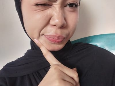 A close-up, waist-up shot shows a young woman winking and sticking out her tongue slightly. She is wearing a black hijab that covers her head and neck. Her right eye is open, and her left eye is closed in a wink. Her mouth is pursed and her tongue is visible between her lips. Her right index finger is touching her chin. The background is a plain, light-colored wall with a turquoise object on the right side. The image appears to be taken indoors in Jeppee, Indonesia.