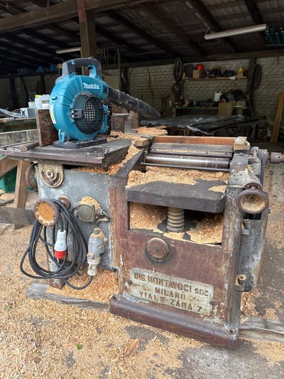 The image shows a woodworking machine, possibly a planer or thicknesser, covered in sawdust in a cluttered workshop environment. The machine has a metal plate with the inscription "ING. MONTAVOCI SOC. MILANO VIALE ZARA 7." A blue Makita blower is positioned next to the machine. Various cables and electrical components are visible near the machine's base. The setting is a workshop filled with tools and equipment. No people are visible. The specific location is likely not Otwock, Poland given that the machine is manufactured in Milano Italy.