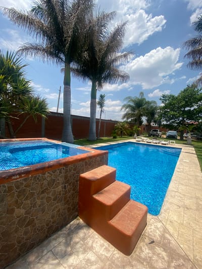 The image depicts a swimming pool and a hot tub in an outdoor setting. The swimming pool is rectangular and filled with blue water, with steps leading into it. Next to the pool is a smaller, square hot tub, also filled with blue water. The surrounding area is paved with light-colored stones. Palm trees are visible in the background, along with a brick wall and other vegetation. The sky is blue with scattered clouds. Several chairs and a white SUV are visible in the background. The location is San Francisco Lachigoló, Mexico.