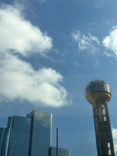 The image presents a striking upward view of an urban skyline against a clear blue sky dotted with white clouds. A prominent architectural landmark, identifiable as Dallas's Reunion Tower, occupies the upper right portion of the frame. This iconic tower features a large, spherical observation deck, encased in a distinctive geodesic grid structure, perched atop a sturdy, concrete column.
Below and to the right of the tower, a modern, glass-clad skyscraper rises into the sky. This building exhibits a stepped architectural design, with multiple reflective surfaces that shimmer under the sunlight. A slender mast or antenna protrudes from its highest point. The perspective is from directly below, emphasizing the towering height of both structures as they reach towards the heavens.
The sky is a vibrant blue, indicating daytime conditions, with scattered cumulus clouds providing texture and contrast. The clouds are bright white, suggesting good weather. No people or specific activities are visible, allowing the architecture and the sky to be the primary subjects. The overall scene captures the grandeur of urban development in Dallas, United States, under a bright, clear sky. No text is visible in the image.