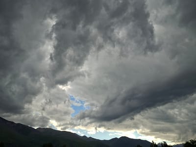 The media file shows a dramatic sky filled with dark, ominous storm clouds. These clouds dominate the upper portion of the frame, with patches of bright blue sky peeking through in a few areas. Below the turbulent sky, the silhouettes of dark, rolling mountains stretch across the horizon. The mountains are covered in green vegetation, suggesting a natural, outdoor setting. The lighting indicates it is either early morning or late afternoon, with the sun partially obscured by the clouds. The overall mood is somber and dramatic, evoking a sense of approaching weather. There are no people or man-made objects clearly visible, except for what appear to be power lines and silhouetted trees in the far distance at the bottom right. The location is identified as Toguz-Bulak, Kyrgyzstan.