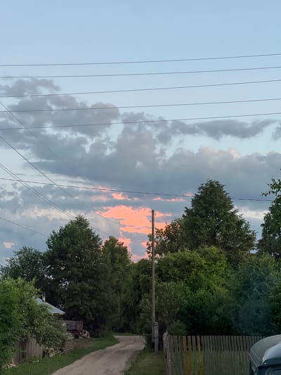The image shows a rural road in the village of Pigozino, Russia, leading towards a dense line of trees. A power line with multiple wires stretches across the upper portion of the frame. A small, rustic house with a metal roof and a chimney is visible on the left side of the road, partially obscured by foliage. A wooden fence runs along the right side of the road, and a utility pole stands near the left edge. In the sky, a band of soft pink and orange clouds is visible through gaps in darker grey clouds, indicating either sunrise or sunset. A few small, pinkish dots, possibly distant kites or balloons, are visible in the upper left quadrant of the sky. The overall scene depicts a quiet, natural environment.