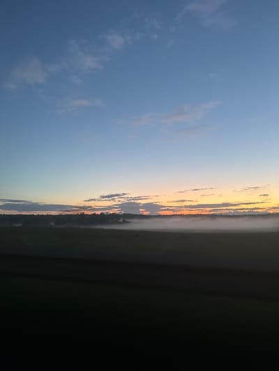 The image depicts a serene, misty landscape at dawn or dusk, likely in the city of Егорье, Russia. The sky transitions from a deep blue at the top to a soft gradient of orange and yellow near the horizon, indicating the sun is either rising or setting. A thin crescent moon is visible in the upper right quadrant of the sky. Wispy clouds are scattered across the sky, adding texture.
Below the sky, a dense band of white mist blankets the horizon, obscuring the base of a treeline that stretches horizontally across the image. The treeline appears as a dark, silhouetted mass against the lighter sky. In the foreground, a dark, unidentifiable area occupies the bottom portion of the frame, possibly a field or road. There are no people, animals, or specific structures clearly visible in the scene. The overall atmosphere is tranquil and natural.